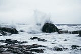 Rock formations in an ocean, white foamy water in foreground, water spraying high against a rock in the background.