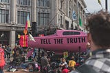 In front of an office building protestors sit in front of a pink water vessel with ‘tell the truth’ in large black letters.