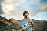 Image of a man with a beanie hat, glasses, and t-shirt pondering thoughts. Background are mountains and semi-cloudy sky, blurred out.