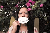 a woman with dark hair covering her mouth with a piece of paper that has a smile drawn on it. A pink rose bush is behind her.