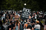 A man standing in a crowd protesting at a park holds up a black sign with white text that reads: Fuck special treatment for cops