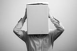It’s a black and white picture. There’s a man standing in front of a plain wall. You see him from the torso up. He’s wearing a striped dress shirt. He’s holding a box over his entire head until his shoulders. You cannot see his head, face or neck.