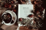 A book is opened to the title page, which reads “Pride and Prejudice, Jane Austen.” Dried roses are scattered over the book and a teacup and saucer sitting next to it.