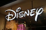 A brightly lit Disney logo sign hangs above a store entrance, featuring the iconic stylized script in white with gold edges.