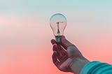 hand outstretched, holding unlit lightbulb among a colorful sunset