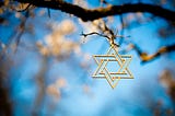 A gold star of david hangs from the branch of a tree against the background of a blue sky and white blossoms