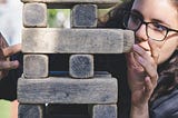 A bespectacled woman carefully adjusts a block in a Jenga-like layer of blocks.