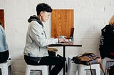 Person sits at table in a coffeeshop while working on their computer.