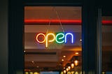 A neon sign hangs in a window, saying “open” with each letter a different color, including red, yellow, green, and blue.