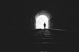 A photo of someone walking out of a dark tunnel into the light of day.