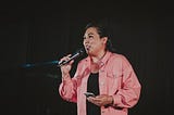 A woman against a black background in a business-casual outfit with a pale pink shirt speaks into a handheld microphone while holding her phone in her other hand.