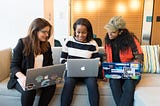 Three women sit on a couch in an office while using laptops.