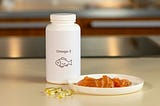 An image of a small container with the label ‘Omega-3 and a drawing of a small fish’, placed next to a bowl containing raw fish, and a handful of insulin, all on a neat table-top.