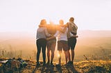 photo of four people standing with their arms around each other’s shoulders, looking away from the camera at rolling hills.