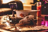 A witch’s workspace, containing a red candle, tarot cards spread, a chalice, a small animal skull, and assorted herbs.