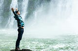 Male standing at the side of a waterfall wearing a cap, sporty clothes, arms up in the air, he looks happy.