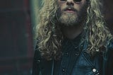 A young man with long curly hair stares directly at the camera. He is wearing dark glasses with round lenses and a black leather jacket.