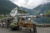 A retired couple sitting on a bench watching a cruise ship by the mountains.