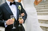 Image of a groom holding two glasses of champagne with the bride standing next to him. Faces not pictured.
