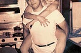 A sepia-toned, black and white photograph of a smiling woman with a dark 1950s hairstyle and bare legs, being carried piggy-back style by a man with dark hair and a white t-shirt. She beams broadly and he grins mischeviously.