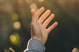 Person wearing a ring on their middle finger, with a hand stretched up to a sunny sky