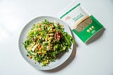 a bag of freekeh seeds next to a plate of salad with veggies and grain, the grain is freekeh.