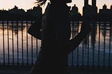 A close up silhouette of a woman with a ponytail runing beside a lake with a city skyline in the background. The sun is setting.