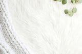 edge of white knit blanket next to a furry white blanket a sprig of green leafs