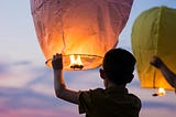 The image depicts a young boy from behind as he is about to release a large, glowing paper lantern into the evening sky. The lantern, illuminated warmly from within, casts a soft orange light on the boy’s hands and the side of his face. He wears a casual short-sleeved shirt and stands against a backdrop of a serene, dusky sky that gradients beautifully from deep blue to shades of purple and pink. To his right, another yellow lantern is also being released.