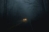 A picture showing a car driving through a very foggy forest at night, only headlights visible