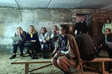 A group of people wearing coats and scarves and sitting on benches while holding a meeting in an underground bunker in Ukraine.