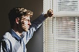 man looking out a window with blinds, thinking