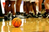 A basketball sits on a school gym floor among an ankle shot of girls in bym shorts.