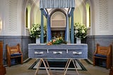 Photo of a blue-grey casket on wooden sawhorses, in front of a blue, curtained archway, set up in an alcove. There are flowers on top of the casket and behind the curtained structure. The walls behind are white with ornate designs on the upper part of the wall, and blue-grey on the lower part of the walls to the wainscotting. There’s a small, wooden bench with blue, padded seat on either side of the image.