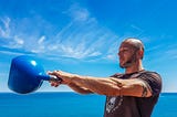 A man helps maintain his weight by swinging  a blue kettlebell. The ocean is in the background.