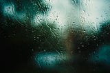 Photograph of a blurred background of light and darkness, seen through a rainy window.