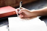 A notebook on a table. Someone’s hand is visible. They are writing.