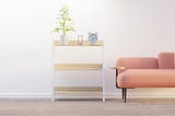 A living room showing a sofa and a shelf that holds an alarm clock, an hour-glass and a plant.