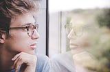 A young person with glasses looking from the window, putting their hand under their chin, and thinking.