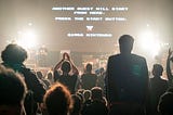 Facing a stage with a rock band on it, three people stand to clap while many more are seated. On the screne above the performers are the words, “Another Quest will start from here Press the start button.”