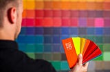 Someone facing away from the camera holding a fan of colour swatches. In front of them, away from the viewer, the wall is covered with out-of-focus squares of many colours.