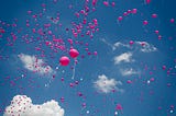 Many pink balloons flying towards a very blue sky, with some clouds.