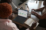 A group of three people sitting around a wooden table with laptops. One person is working on a laptop displaying a calendar or scheduling tool in an Excel-like interface. The individual is holding a coffee cup while taking notes. This image illustrates a collaborative working environment where team members are discussing or reviewing data on their laptops.