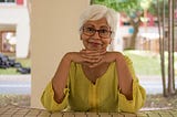 Asian woman with white hair and glasses, hands folded under her chin, wearing a bright yellow shirt