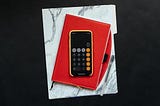 A mobile phone has the calculator app open which sits on top of a red notebook and white marble folder.