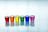 Six shot glasses of alcohol neatly lined up in a row. Each alcohol has a different color and their line-up makes a rainbow color combination.