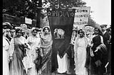 Image description: A black and white photo of Indian suffragettes at the 1911 Coronation Procession holding a triangular flag with the word India and an elephant beneath.