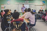 A teaching is standing in front of a class of attentive young students.