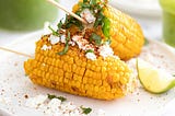 corn on the cob broken in half then put on a skewer for easy eating, served with a sprinkle of cheese, spices, fresh herbs and a wedge of lime on a white plate