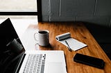 An open laptop is sat upon a wooden desk. Scattered around the laptop is a mug of coffee, a notepad and pen, and a smartphone.